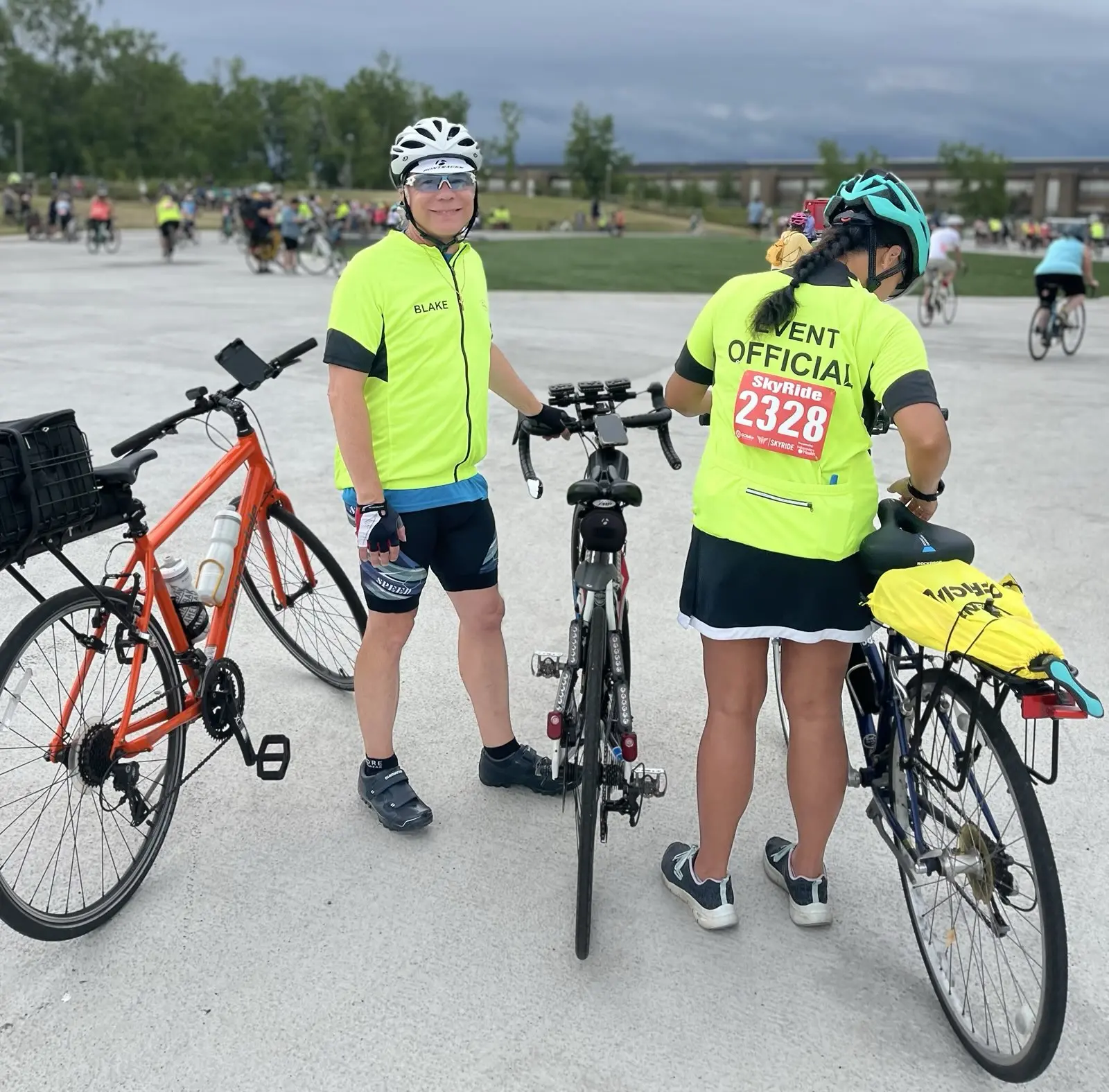Two of our event marshals during a ride, standing next to their bikes, one turned to the camera and smiling, the other inspecting her bike